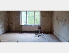 Ein leerer, heruntergekommener Raum mit schmutzigen Wänden, freiliegendem Boden, einem großen Fenster mit Blick auf grüne Vegetation und einem Heizkörper darunter. In der Mitte steht ein einzelner Stuhl mit einigen Gegenständen darauf, umgeben von verstreutem Müll.