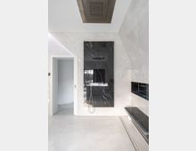 Modernes Badezimmer mit glänzenden weißen Wänden und Bodenfliesen aus Marmoroptik. Eine große, rechteckige schwarze Wandfläche mit einem integriertem Duschkopf und Schlauch ist zentral zu sehen; rechts davon eine schmale eingelegte Ablage und links eine Türöffnung.