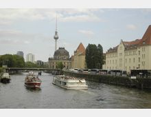 Flusslandschaft in Berlin mit der Spree, auf der Ausflugsboote fahren, im Hintergrund das markante Bode-Museum mit Kuppeldach und der Fernsehturm. Auf der rechten Seite sind historische Gebäude und mehrere geparkte Reisebusse zu sehen.