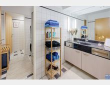 Helles Badezimmer mit weißen Wandfliesen, einem Waschbecken auf einer dunklen Arbeitsplatte, Holzunterschränken und einem Spiegel mit integrierter Beleuchtung. Daneben steht ein offenes Holzregal mit Handtüchern und Pflegeprodukten, und im Hintergrund ist ein Zugang zu einem separaten Raum sichtbar.