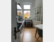 Helle Küche mit Holzboden und weißen Möbeln: links eine Küchenzeile mit Spüle, Schränken und Arbeitsfläche, rechts ein weißer Vitrinenschrank und ein Esstisch mit Stuhl. Ein großes Fenster mit Blick auf Bäume in Herbstfarben dominiert den Hintergrund.