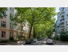 Baumbestandene Wohnstraße mit mehrstöckigen Altbauten auf beiden Seiten, teils mit Balkonen und Blumenschmuck. Geparkte Autos säumen die ruhige Straße, die von großen, belaubten Bäumen überspannt wird.