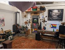 Ein rustikaler Raum mit weiß gestrichenen Backsteinwänden, einem Holzofen in der Ecke und einer Tür mit einem runden Fenster, umgeben von dekorativen Elementen. Der Raum enthält eine Vielzahl von Gegenständen wie einen Einkaufswagen mit Pflanzen, Holzmöbel, bunte Poster, Kunstwerke und handwerkliche Dekorationen, auf einem roten Teppichboden.