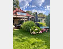 Einfamilienhaus mit rotem Ziegeldach und überdachter Terrasse, davor eine gepflegte Rasenfläche. Im Vordergrund liegen ein bepflanztes Beet mit Ziergräsern und blühenden Hortensien sowie ein kleiner Poolbereich mit Sonnenschirmen.