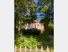 Eine rosa Villa mit weißen Fensterrahmen, teilweise verdeckt von hohen Bäumen und üppiger Vegetation, steht hinter einem schmiedeeisernen Zaun. Der Vorgarten ist mit grünen Pflanzen und Blumen bewachsen, und im Hintergrund ist ein klarer blauer Himmel zu sehen.