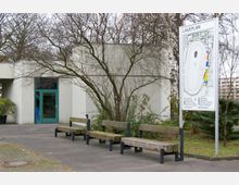 Ein kleiner Außenbereich mit mehreren Holzbänken auf einer betonierten Fläche, im Hintergrund ein modernes, flaches Gebäude mit hellem Verputz und einem Eingang mit grüner Tür. Rechts im Bild steht ein großer Lageplan, und kahles Geäst sowie Büsche umrahmen die Szene.