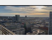 Luftaufnahme einer städtischen Landschaft mit zahlreichen Gebäuden unterschiedlicher Höhe, darunter moderne Hochhäuser und farbige Altbauten. Im Hintergrund ein weitläufiger Blick über die Stadt mit einem klaren Himmel und vereinzelten Wolken bei Sonnenuntergang.