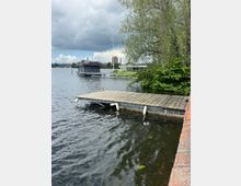 Holzsteg am Ufer eines Sees mit dunkler Wasseroberfläche und Bojen, rechts von überhängenden Bäumen und Sträuchern eingerahmt. Im Hintergrund liegt ein modernes Hausboot am Wasser, dahinter weitere Gebäude unter bewölktem Himmel.