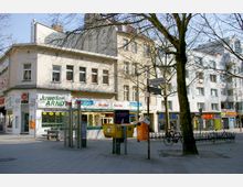 Straßenecke mit mehrstöckigen Wohn- und Geschäftsgebäuden, darunter Läden wie ein Juweliergeschäft und ein Reformhaus. Im Vordergrund stehen Briefkästen, eine orangene Altkleiderbox, Telefonzellen und ein Baum; Straßenschilder weisen auf die Schlossstraße und Deimelstraße hin.