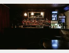 Dimly lit bar interior with a long counter, shelves of liquor bottles, and three hanging pendant lights reflected in a large wall mirror. Red curtains frame the back wall, with a chalkboard menu on the left and illuminated beverage refrigerators on the right.
