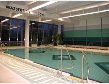 Innenansicht eines Schwimmbads mit einem rechteckigen Becken, einer angegebenen Wassertiefe von 1,30 m. Große Fenster an der Wand lassen Blick nach draußen zu, während grüne Pflanzen und eine blau-grüne Wandgestaltung die Umgebung ergänzen.