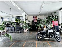 Geräumiger, lichtdurchfluteter Innenraum mit industriellem Stil, ausgestattet mit einer Bar, Stühlen, zahlreichen Zimmerpflanzen, einem großen Tisch und einer schwarzen Motorradmaschine im Vordergrund. Im Hintergrund sind Regale, ein roter Schrank und moderne Kunst an der Wand sichtbar.