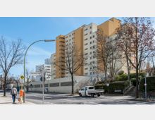Mehrstöckiges Wohngebäude mit gelben und weißen Fassaden, umgeben von Bäumen und Gehwegen. Im Vordergrund sind eine Straßenkreuzung, ein Straßenschild mit der Aufschrift "Domnauer Straße", eine Bushaltestelle und ein parkender Kleintransporter zu sehen.