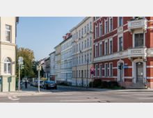 Straßenszene mit mehrstöckigen Wohnhäusern im Altbaustil, teils mit roten Klinkerfassaden und weißen Stuckverzierungen. Im Vordergrund sind eine am Gehweg geparkte Autoschlange, Verkehrszeichen und eine Ampel sichtbar, im Hintergrund Bäume und ein blauer Himmel.