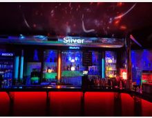 Innenansicht einer Bar mit beleuchteter Theke und Regalen voller Flaschen und Gläser, dominiert von blauem und rotem Neonlicht. Über der Bar hängt ein leuchtendes Schild „Silver Event Location“, an der Decke sind farbige Lichtprojektionen zu sehen.
