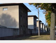 Reihe von einfachen, mehrstöckigen Wohngebäuden mit flachen Dächern und verputzten Fassaden, entlang einer Straße mit Bäumen und Strommasten am Rand. Der Boden ist mit Herbstlaub bedeckt, und die Szene wirkt ruhig und sonnenbeschienen.