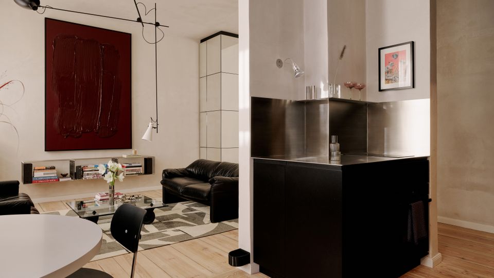 Offener Wohn- und Essbereich mit hellem Holzboden, schwarzer Küchenzeile mit Edelstahlrückwand und weiß verputzten Wänden. Im Hintergrund stehen schwarze Ledersofas und ein Glastisch auf einem gemusterten Teppich, darüber eine moderne Pendelleuchte und ein großes, dunkelrotes Wandbild.