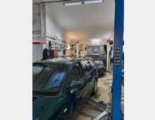 Innenansicht einer Autowerkstatt mit zwei geparkten Autos, eines dunkelgrün und eines silbern, auf Hebebühnen. Die Wände sind mit Nummernschildern und Werkzeug dekoriert, und die Beleuchtung besteht aus Deckenlampen.