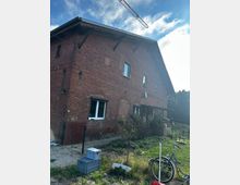 Backsteinhaus auf einem Grundstück mit Garten, umgeben von niedrigen Zäunen. Im Vordergrund steht ein Fahrrad, und im Hintergrund ragt ein Baukran in den Himmel.