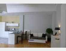 Wohnzimmer mit integriertem Küchenbereich in modernem Design. Zu sehen sind eine weiße Küche mit Arbeitsfläche, ein weißes Sofa mit schwarzen Kissen, ein Couchtisch, ein Topfpflanzenarrangement und ein Holzfußboden im Fischgrätmuster.