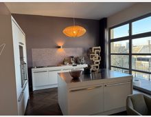 Modern open-plan kitchen with white cabinets, a central island with a dark countertop, and a pendant light above. Large floor-to-ceiling windows and sliding door open to a balcony, bringing in daylight and showing nearby rooftops.
