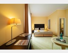 Ein Hotelzimmer mit gelb gestrichenen Wänden und einem Doppelbett mit dunklem Kopfteil, flankiert von zwei Nachttischen mit Lampen. Im Vordergrund steht ein Schreibtisch mit einer Tischlampe und Magazinen, sowie ein runder Beistelltisch mit Wasserflasche und Gläsern; daneben ist ein Badezimmer sichtbar.