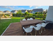 Holzterrasse mit großem rechteckigem Esstisch und mehreren weißen Stühlen, neben einer Außenfeuerstelle. Dahinter ein gepflegter Garten mit Rasen, Hecken, Hochbeet und Hängematte unter einem Sonnensegel; im Hintergrund stehen moderne Wohnhäuser.