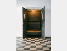 Alter Lastenaufzug mit offenen schwarzen Türen, einem dunklen Innenraum und einem beleuchteten Deckenbereich; der Boden davor ist mit schwarz-weiß karierten Fliesen ausgelegt.