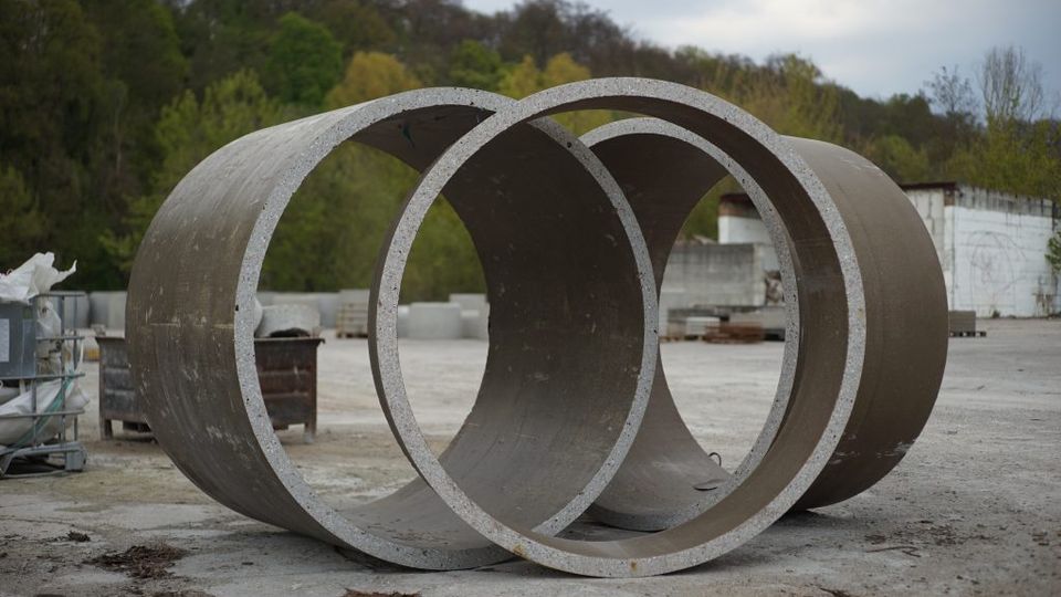 Zwei große Betonringe liegen auf einem offenen, betonierten Lagerplatz. Im Hintergrund sind grüne Bäume, eine weiße Wand und gestapelte Baumaterialien zu sehen.