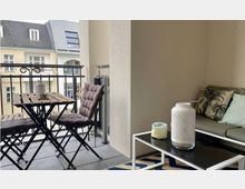 Balkon mit Sitzbereich, bestehend aus einem kleinen Holztisch mit zwei gepolsterten Klappstühlen und einer modernen Couch mit Kissen. Im Vordergrund steht ein niedriger Glastisch mit Dekoration, im Hintergrund ist ein schmiedeeisernes Geländer und Wohnhäuser sichtbar.