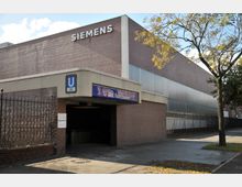 Eingang zur U-Bahn-Station "Rohrdamm" der Linie U7 in Berlin, flankiert von einem Ziegelgebäude mit dem Schriftzug "Siemens" und einem Zaun. Im Vordergrund sind ein baumbestandener Gehweg und ein Hinweisschild sichtbar.