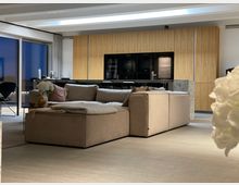 Modernes Wohnzimmer mit offener Küche, beige Ecksofa im Vordergrund, dahinter Holzschränke und schwarze Einbaugeräte. Große Fenster auf der linken Seite lassen Blick nach draußen zu.