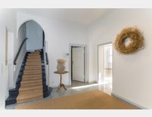 Heller Eingangsbereich mit schwarzer Treppe, beige Teppichläufer auf Marmorboden, einem runden Holztisch mit Blumenvase und einem trockenen Kranz an der weißen Wand. Türen führen zu weiteren Räumen, einer davon mit viel Tageslicht.