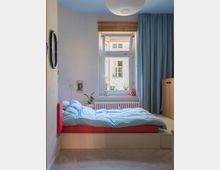 Ein kleines Schlafzimmer mit einem erhöhten Bett, das von hellblauer Bettwäsche bedeckt ist. Im Hintergrund befindet sich ein Fenster mit einem Holzrollo, flankiert von blauen Vorhängen, das Tageslicht in den Raum lässt; eine weiße Papierlampe hängt von der Decke.