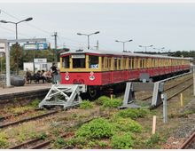 Eine historische rote und gelbe S-Bahn der Linie S3 mit der Zielanzeige "Friedrichstr." steht an einem Endbahnhof in Erkner. Im Hintergrund sind eine Uhr, das Bahnhofsschild sowie wartende Personen auf der Bahnsteigbank zu sehen, umgeben von Vegetation und oberirdischen Stromleitungen.