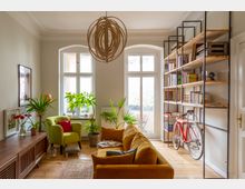 Helles Wohnzimmer mit hohen Decken, Holzfußboden und zwei großen Sprossenfenstern sowie einer Glastür zum Balkon. Rechts steht ein offenes Metall-Holz-Regal mit Büchern und einem angelehnten roten Fahrrad; im Raum sind ein Sofa, ein Sessel, mehrere Zimmerpflanzen und eine große runde Hängelampe.