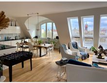 Helles Wohnzimmer mit offener Küche in einer Dachgeschosswohnung, ausgestattet mit Holzfußboden. Große Fenster bieten Blick auf eine städtische Kulisse mit Kirchturm, während der Raum mit einem Esstisch, entspannenden Sesseln, Pflanzen und einem Tischkicker eingerichtet ist.