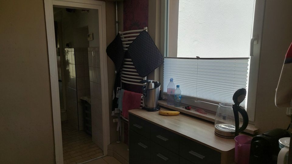 Kleiner Küchenbereich mit Holzarbeitsplatte und Unterschränken neben einem großen Fenster mit halb geschlossenem Plissee. Links führt eine Tür in einen gefliesten Nebenraum, daneben steht ein Wäscheständer mit aufgehängten Textilien; auf der Fensterbank und Arbeitsfläche stehen Wasserflaschen und eine Kaffeemaschine.