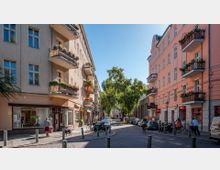 Straßenszene in einem Wohnviertel mit mehrstöckigen Altbauten, links ein beigefarbenes Gebäude mit dekorativen Balkonen, rechts ein rosafarbenes Gebäude. Im Vordergrund befinden sich kleine Geschäfte, Fahrräder und geparkte Autos, während ein großer Baum die Mitte der Straße beschattet.