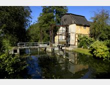 Historisches Wasserkraftwerk mit gelbem Gebäude und Holzdach, umgeben von Bäumen und Vegetation, mit einem kleinen Wehr und reflektierendem Wasser im Vordergrund.