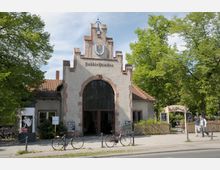 Historisches Gebäude im Stil der Backsteinarchitektur mit einem großen Torbogen und der Beschriftung „Poddhülskiallee“ darüber, flankiert von roten Dachziegeln und Bäumen im Hintergrund; vor dem Eingang stehen Fahrräder, und rechts führt ein Weg zu einem Café-Garten mit einem geschwungenen Torbogen.