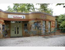 Flachbau aus gelben Klinkersteinen mit einem Schild "Freizeitstätte Koeltzepark" über dem Eingang; die Fassade ist mit Graffiti bedeckt. Im Vordergrund befindet sich ein gepflasterter Weg, umgeben von Bäumen und Sträuchern.