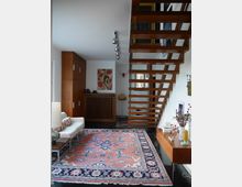 Wohnraum mit modernem Design; eine offene Holztreppe führt nach oben, darunter helle Wände und Möbel. Im Vordergrund liegt ein großer, farbiger Teppich mit einem floralen Muster, begleitet von einem Sofa, einem Sideboard und dekorativen Elementen.