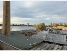 Blick über flache Industriedächer mit einem hohen gemauerten Schornstein im Vordergrund, daneben teils verwitterte Dachflächen mit Oberlichtern. Dahinter liegt ein breites Gewässer mit Uferbäumen und einer Stadt-Silhouette aus Wohn- und Gewerbebauten unter bewölktem Himmel.
