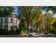 Straßenszene mit einem großen, mehrstöckigen Altbau im Vordergrund, umgeben von dichtem Grün und hohen Bäumen. Entlang der kopfsteingepflasterten Straße parken mehrere Autos, darunter ein Taxi, und die Umgebung wirkt ruhig und von Wohnhäusern geprägt.