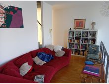 Wohnzimmer mit Parkettboden, einem roten Ecksofa und einem kleinen Beistelltisch. Im Hintergrund steht ein Bücherregal, darüber Bilder und eine Büste; daneben ein gemusterter Sessel. An der Wand hängt ein farbenfrohes Kunstwerk.
