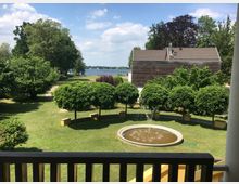 Blick auf einen gepflegten Garten mit rundem Springbrunnen im Vordergrund, umgeben von Bäumen und Rasenflächen. Im Hintergrund befinden sich ein modernes Holzgebäude und ein See, umgeben von weiteren Bäumen.