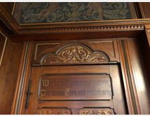 Holzvertäfelte Wand mit kunstvoll geschnitzten Verzierungen und einem oberen Bereich mit gemaltem Ornamentmuster. In die Holzfläche ist ein Schriftzug in deutscher Frakturschrift eingraviert.