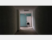 Ein schmaler, schlecht beleuchteter Flur mit grünlichen Wänden und einer weißen Tür am Ende. Vor der geöffneten Tür steht ein einzelner schwarzer Stuhl.