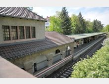 Bahnhof mit einem historischen Gebäude aus hellem Putz und großen Fenstern neben einem Bahnsteig mit grüner Überdachung; im Vordergrund Blumen und Büsche, im Hintergrund Bäume und ein Wohnhaus.